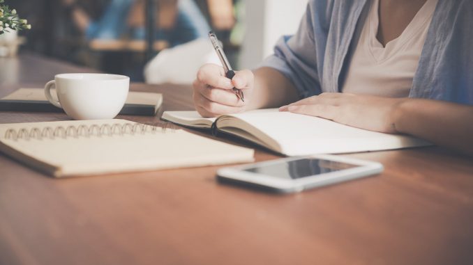 woman writing on a notebook beside teacup and tablet computer